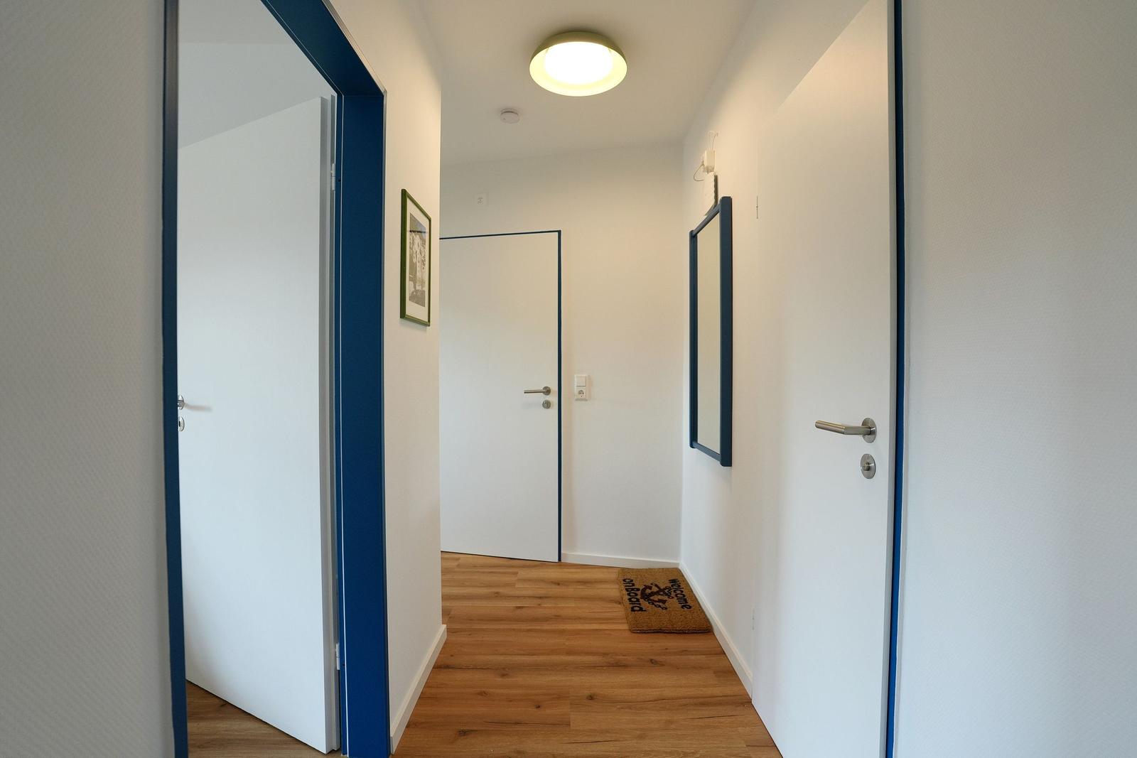 Flur mit Holzboden, weißen Wänden und blauen Türumschlägen. Vordergrund: Tür mit Türmatte.