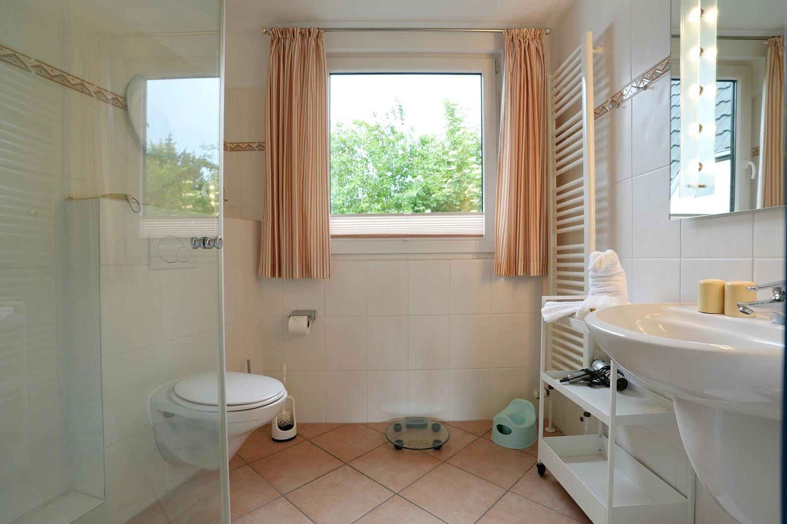Badezimmer mit Dusche, Toilette, Waschbecken und Fenster mit Vorhängen.