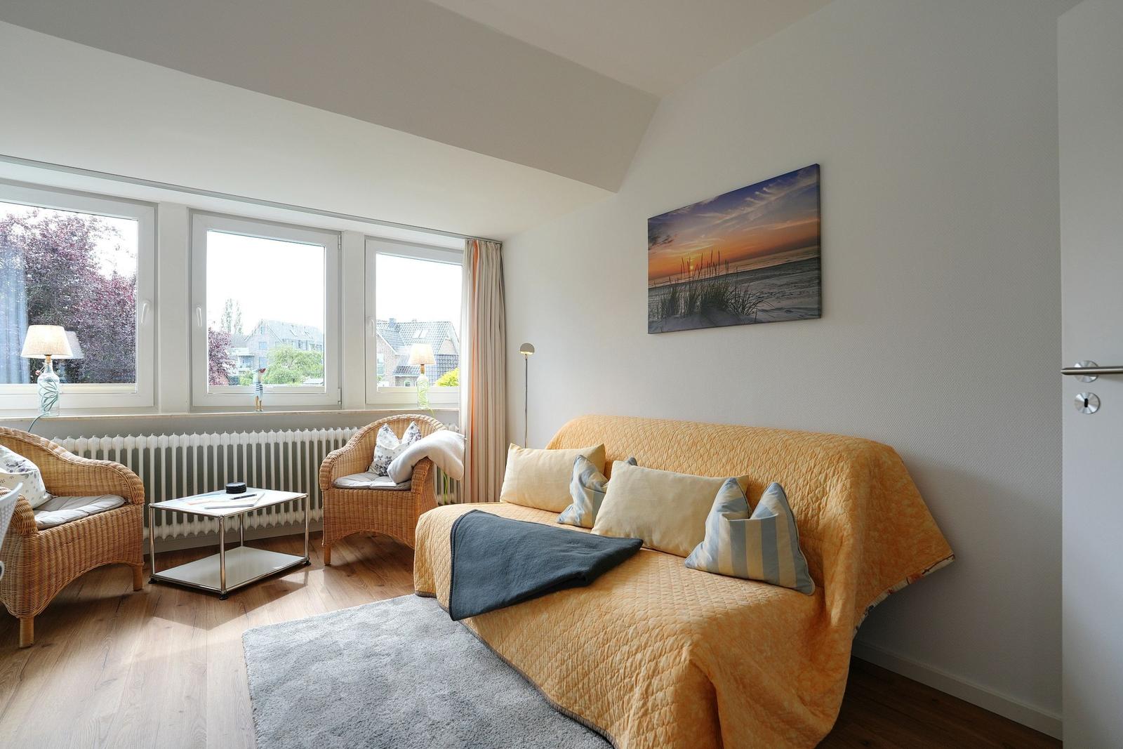 Wohnzimmer mit Sofa, zwei Sesseln und großen Fenstern. Wandbild zeigt Strand bei Sonnenuntergang.
