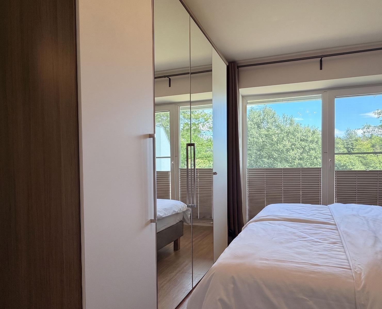 Schlafzimmer mit Doppelbett, Spiegelschrank und Blick auf Balkon mit Bäumen.