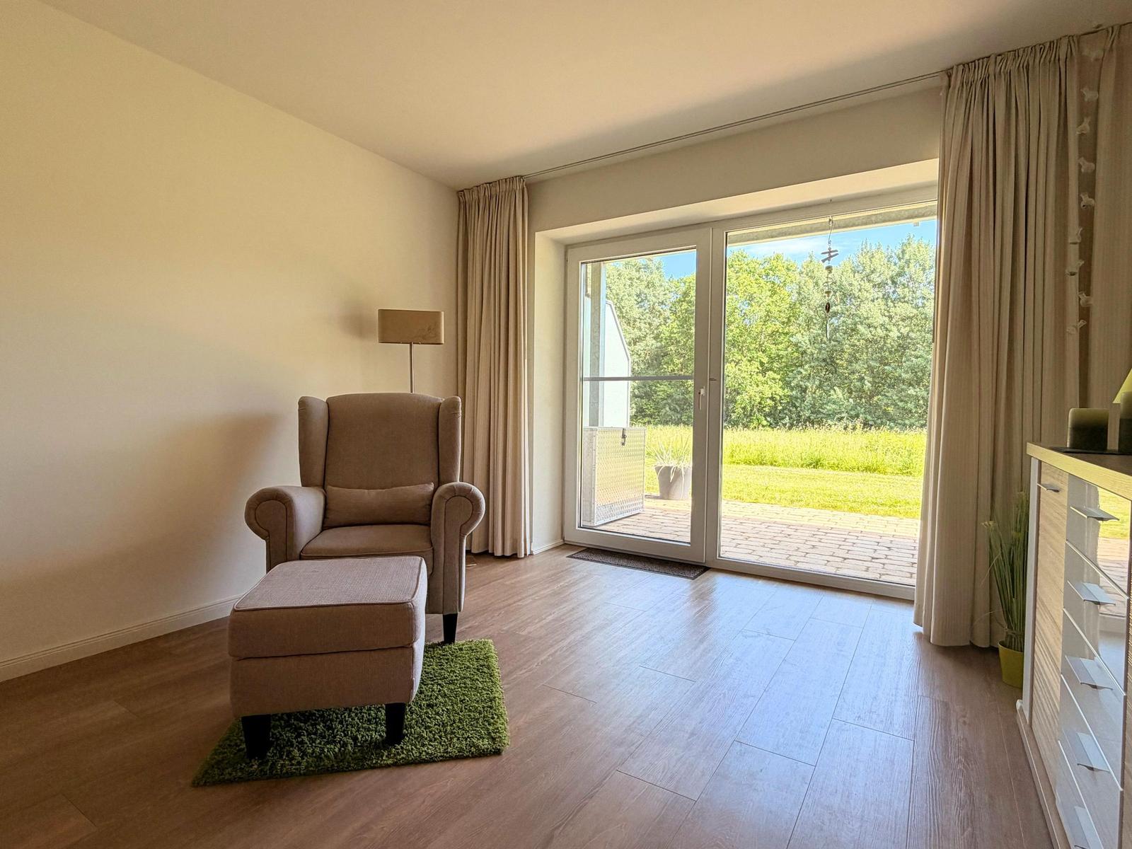 Ein Raum mit Sessel und Fußstütze vor großem Fenster mit Blick auf Garten.