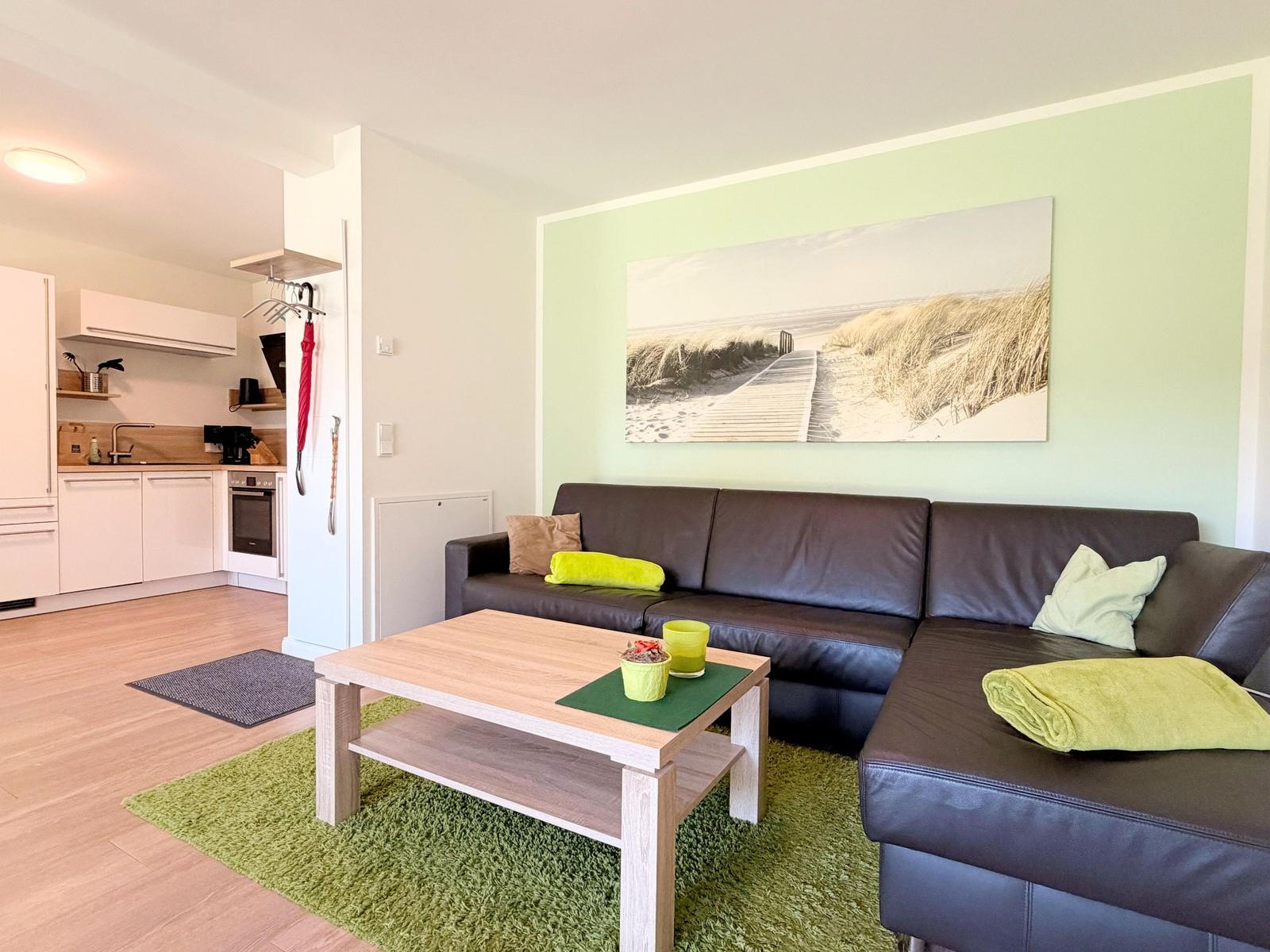 Wohnzimmer mit Sofa, Couchtisch und Küchenbereich. Wandbild zeigt Strand.