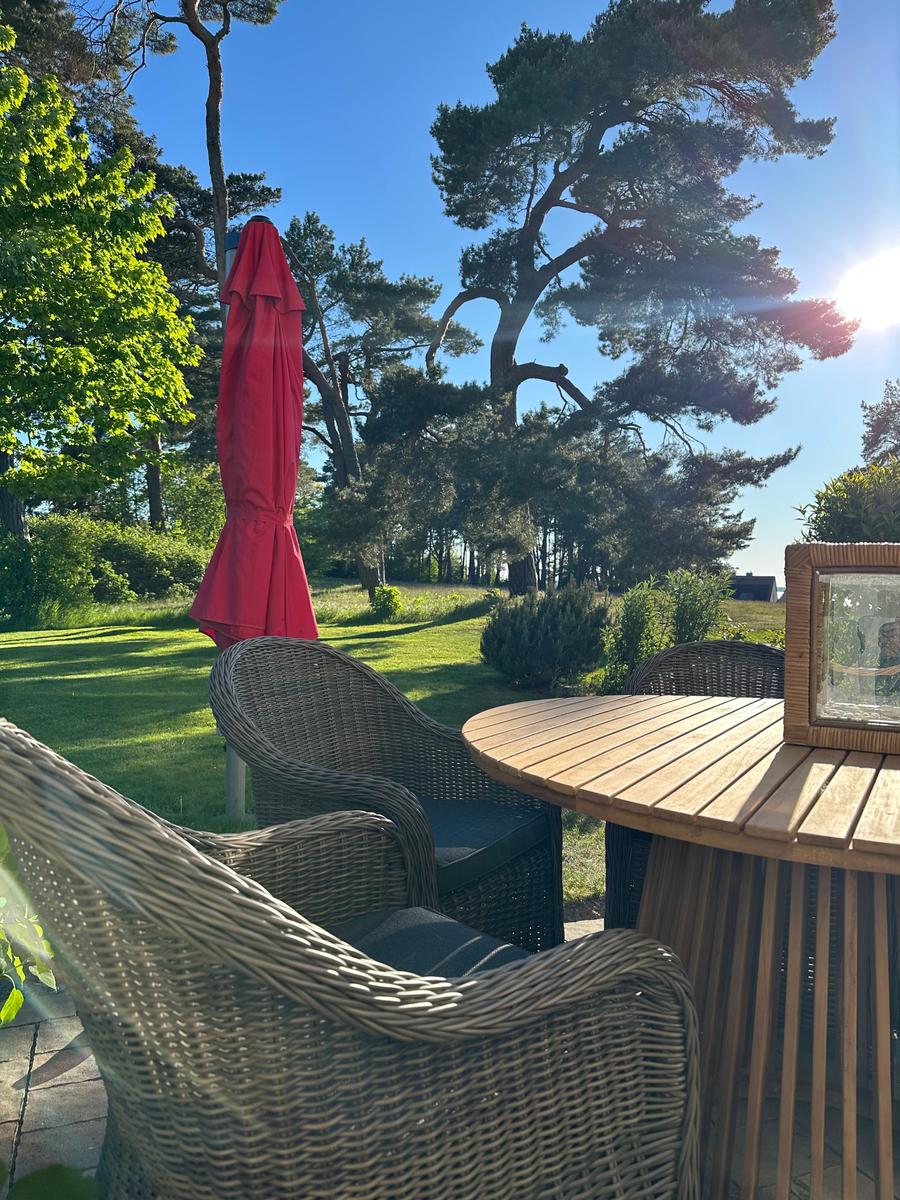 Terrasse mit Rattanmöbeln, Holztisch und rotem Sonnenschirm. Grüner Garten und Bäume im Hintergrund.