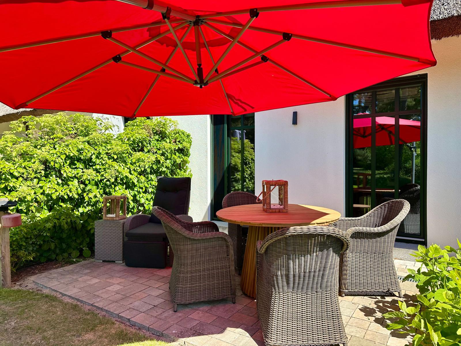 Terrasse mit runden Tisch, vier Stühlen und rotem Sonnenschirm. Hintergrund: grüne Hecke und Fenster.