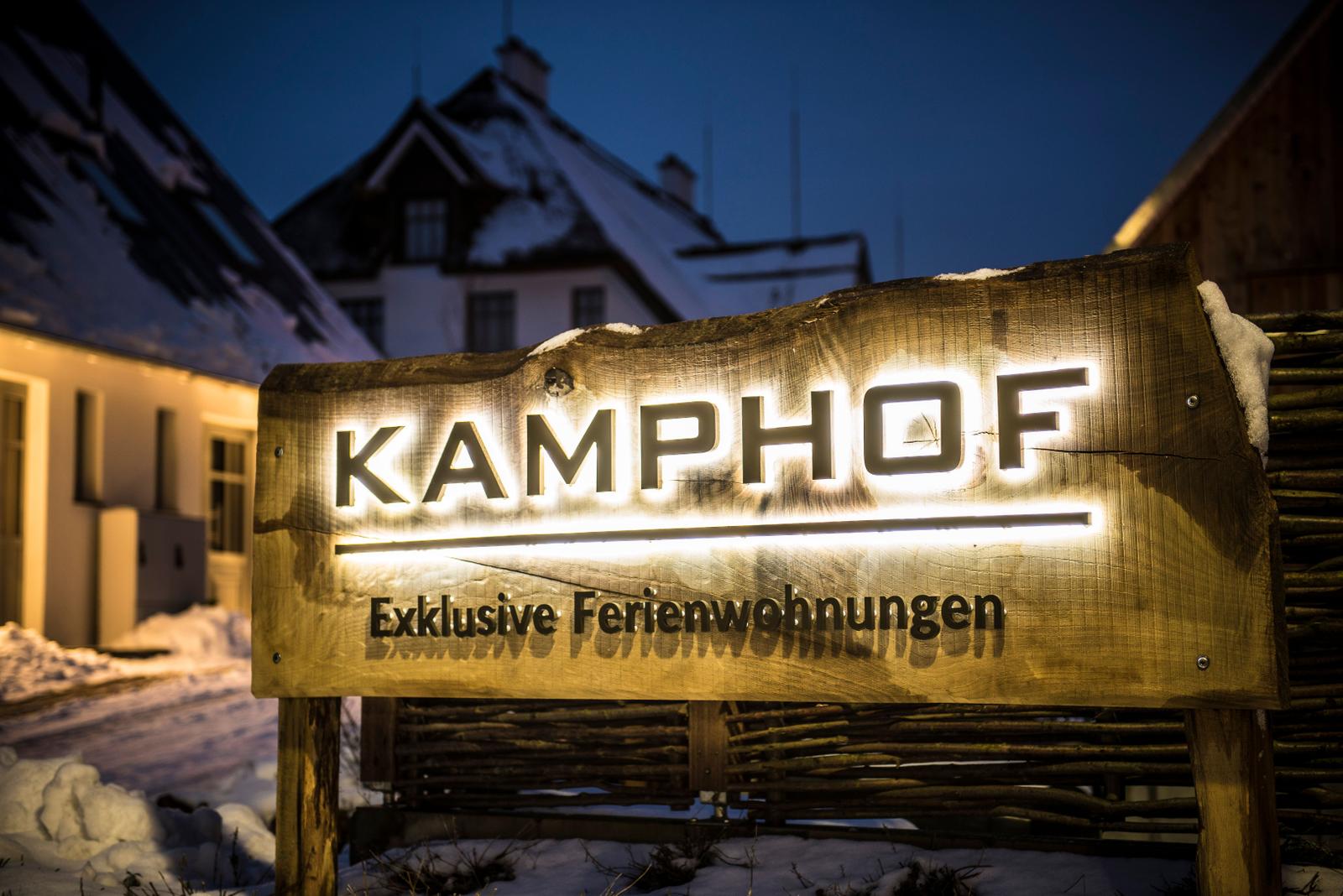 Kamp Hof: Exklusive Ferienwohnungen. Holzschild mit Beleuchtung vor schneebedeckten Gebäuden.