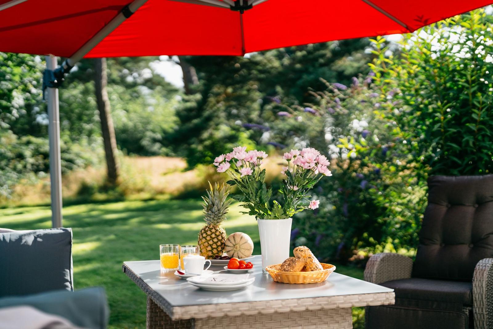 Tisch unter rotem Sonnenschirm mit Obst, Blumen und Kaffee.