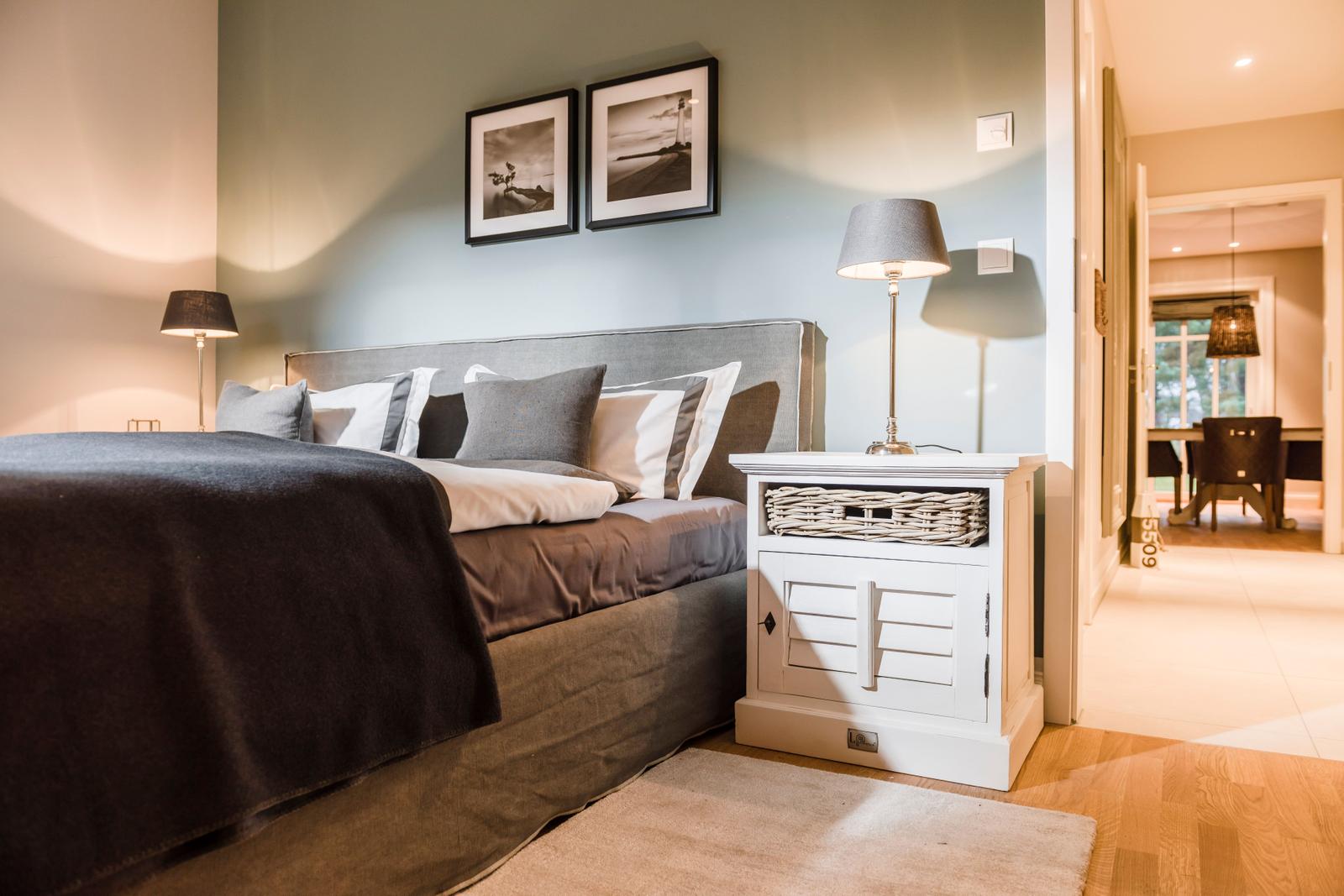 Schlafzimmer mit Bett, Nachttisch und Wandbildern. Durchgang zur Küche sichtbar.
