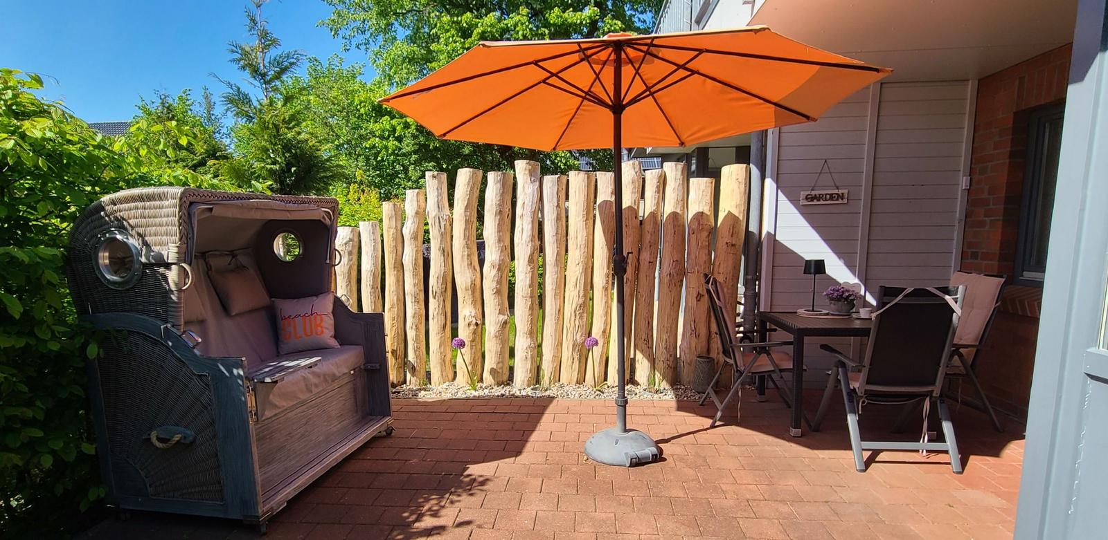 Terrasse mit orangefarbenem Sonnenschirm, Holzzaun und Gartenmöbeln.