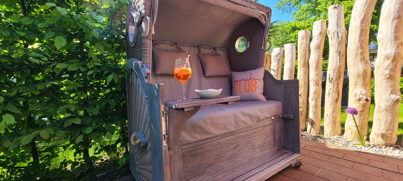 Ein Strandclub-Deckchair mit Getränk und Kissen auf einer Terrasse mit Holzpfosten und Grün.