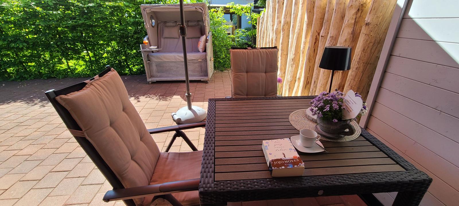 Terrasse mit Tisch, Stühlen und Sonnenschirm. Hintergrund: Holzwand und grüne Hecke.