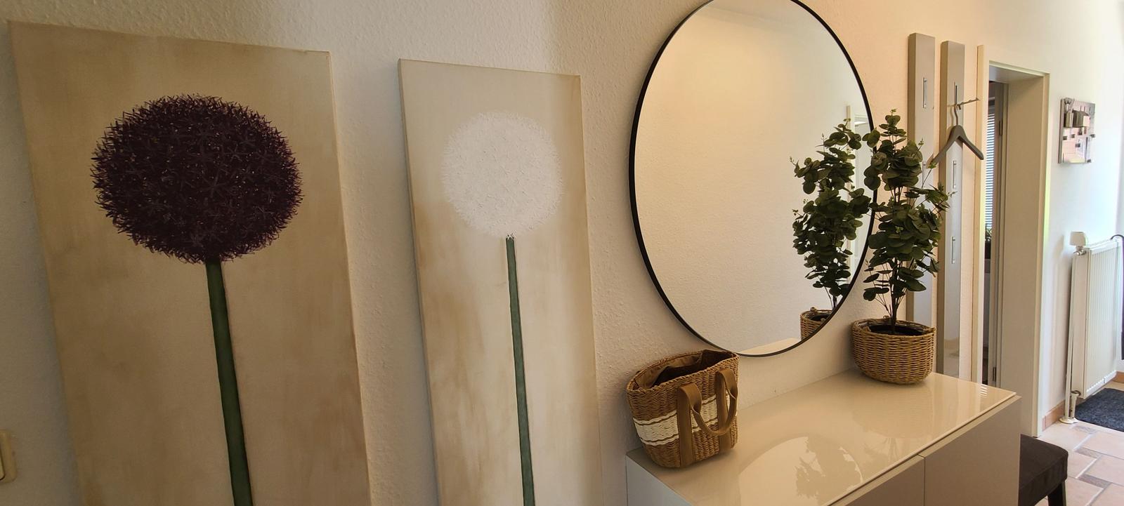 Wand mit zwei großen Bildern und runder Spiegel über Schrank mit Pflanze und Korb.