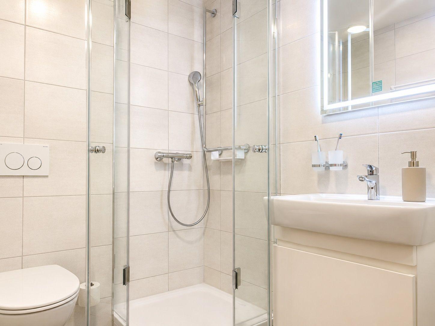 Badezimmer mit Dusche, Waschbecken und Toilette. Fliesenwand und Spiegel mit Beleuchtung.