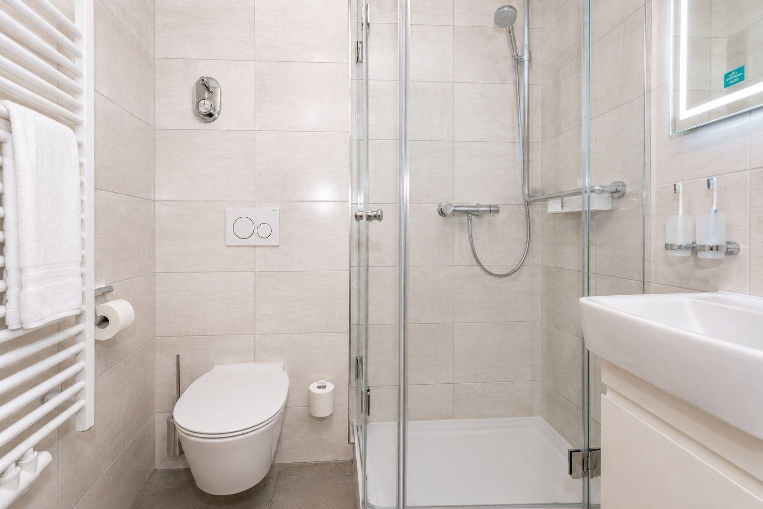 Badezimmer mit Dusche, WC und Waschbecken. Fliesenwand und weißes Sanitär.