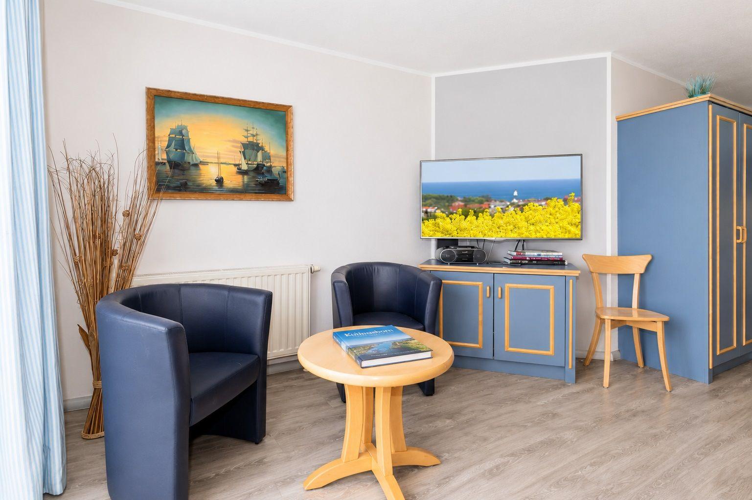 Zimmer mit TV, zwei Sesseln, Tisch und Kleiderschrank. Wandbild mit Schiffen.