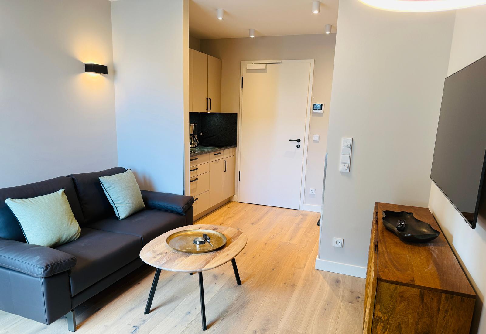 Wohnzimmer mit Sofa, Couchtisch und Küchenbereich. Flur mit Tür und Heizungselement.