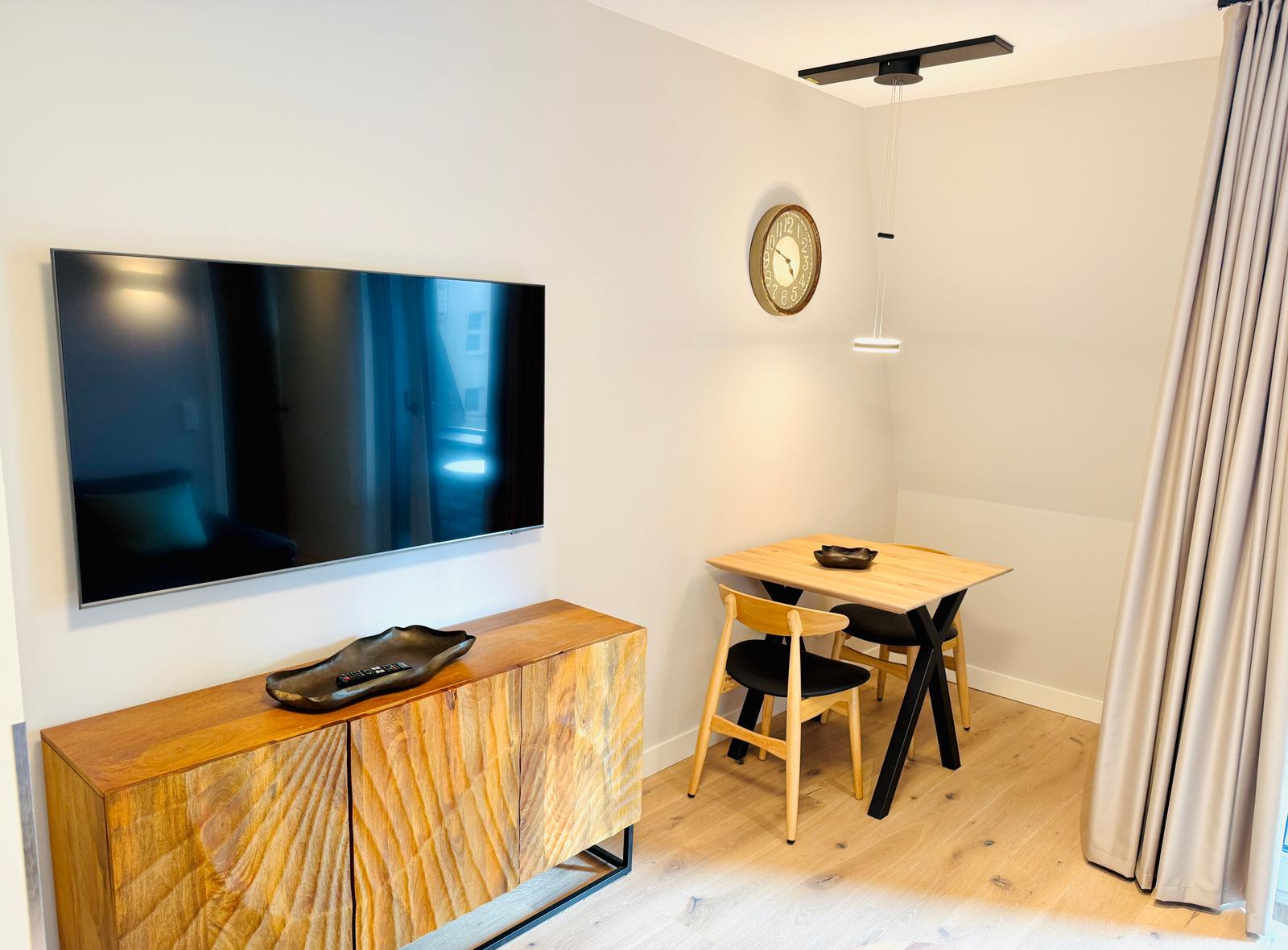 Ein Raum mit Fernseher, Holzschrank, Esstisch und Stühlen sowie einer Wanduhr.
