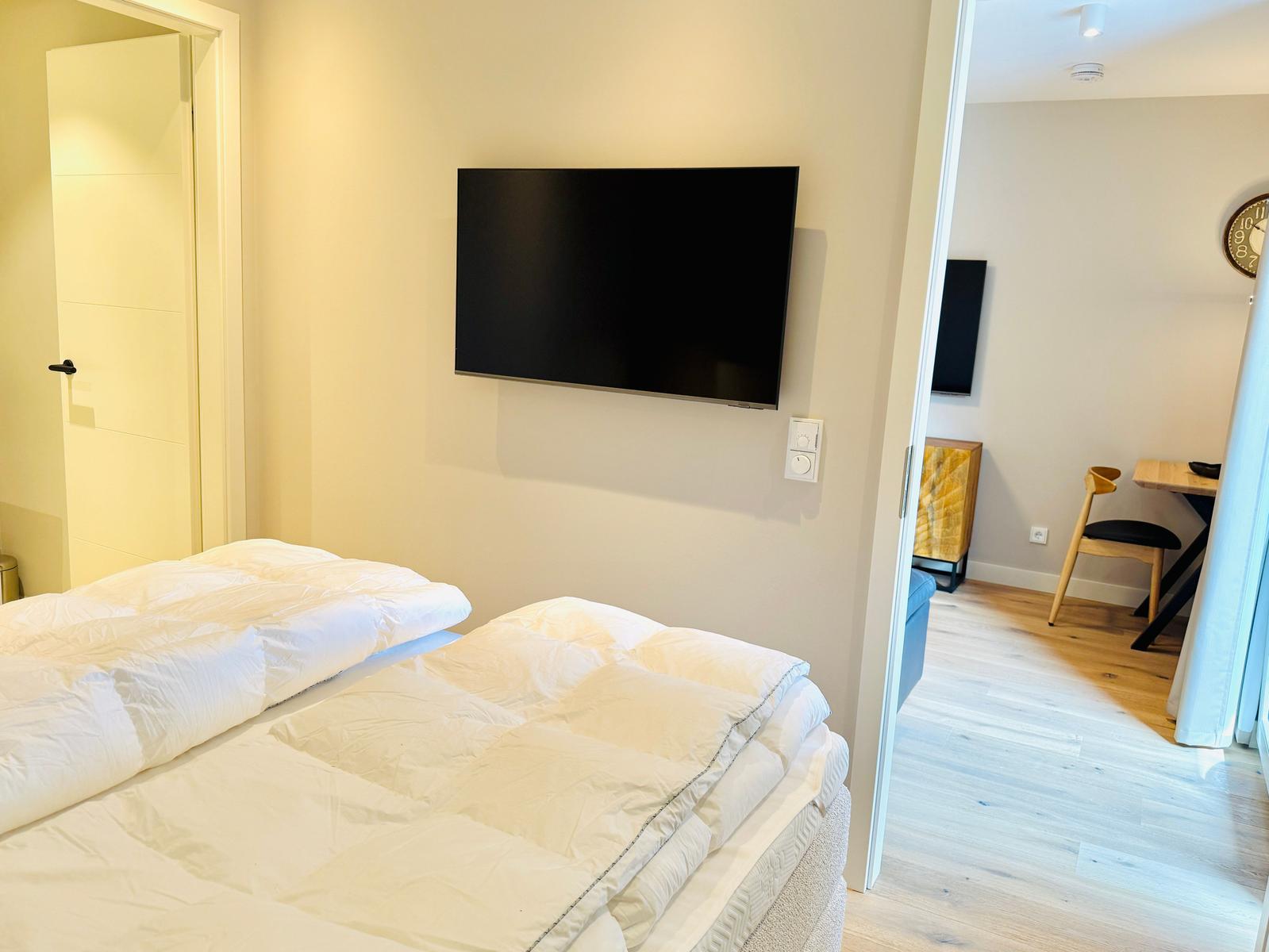 Zimmer mit Bett und Wandfernseher. Durchgang in Wohnbereich mit Tisch und Stuhl.
