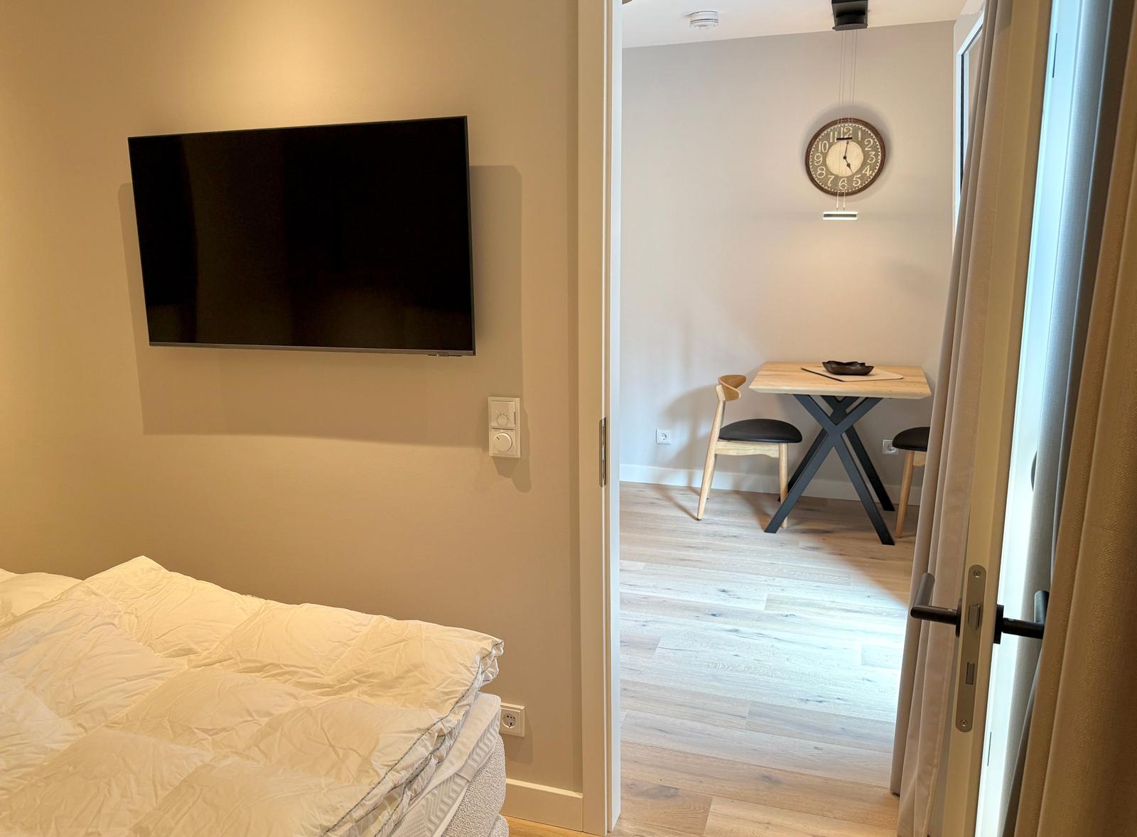 Schlafzimmer mit TV und Blick in Essbereich mit Tisch und Stühlen.