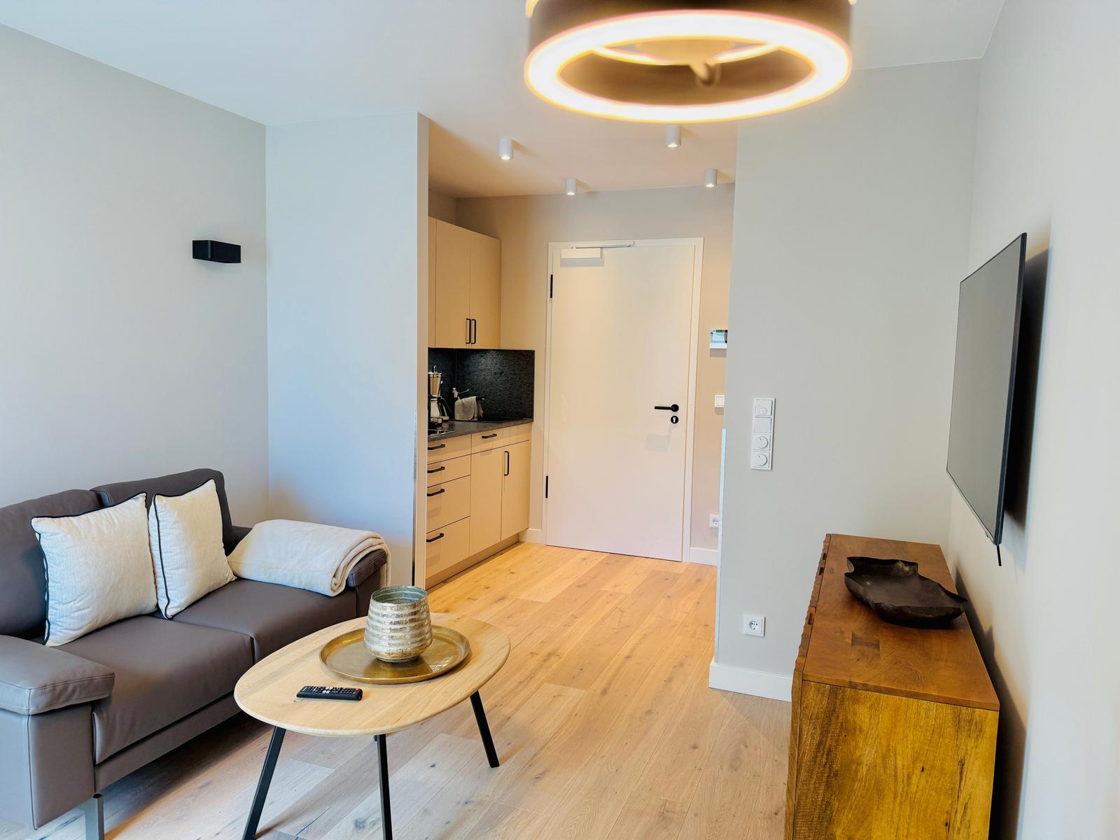 Wohnzimmer mit Sofa, Couchtisch und Fernseher. Küche im Hintergrund.