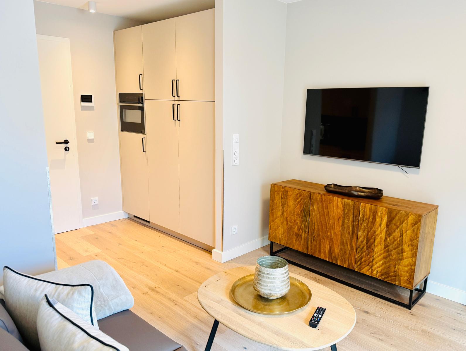Wohnzimmer mit Holzboden, Fernseher und Sideboard. Küchenbereich mit Hochschrank sichtbar.