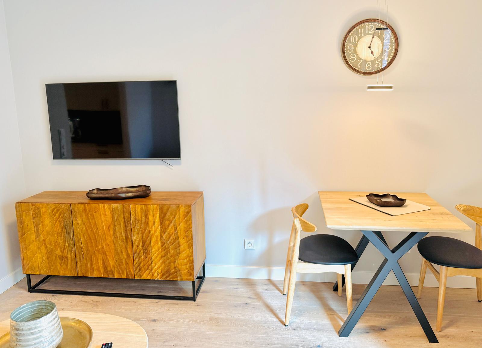 Wohn- und Essbereich mit Holztisch, Stühlen, Fernseher und Wanduhr.