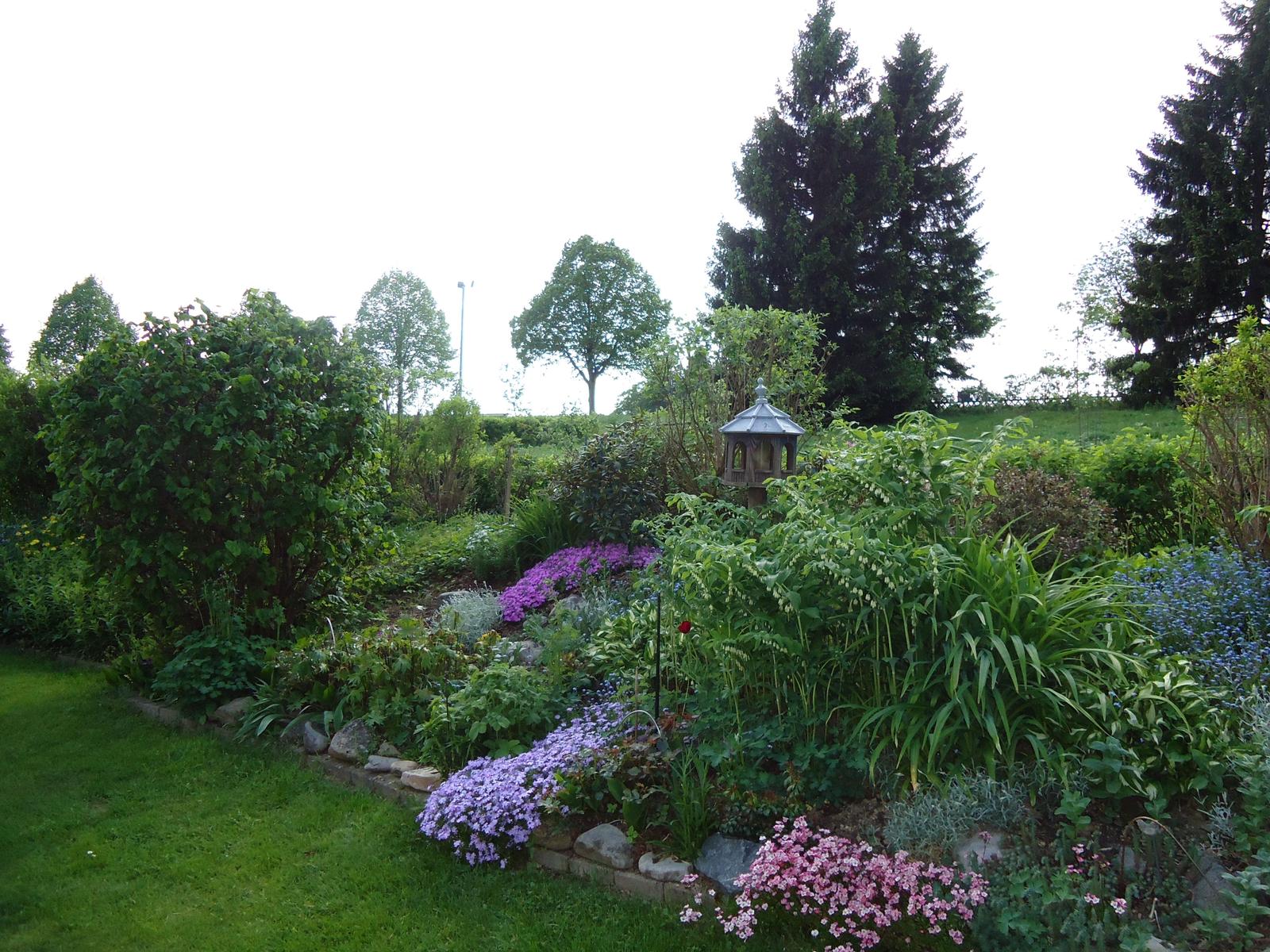 Garten mit Blumenbeeten, Sträuchern und einem Pavillon. Hintergrund mit Bäumen.