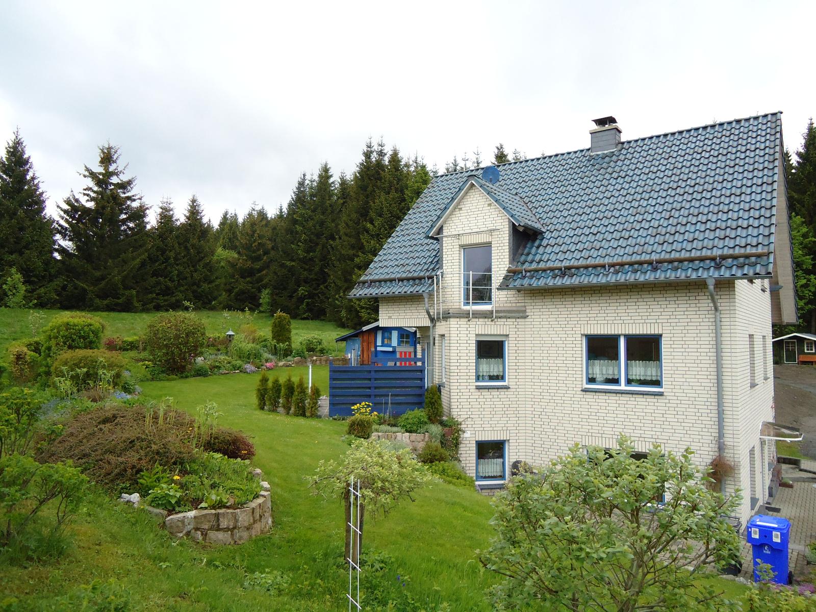 Haus mit Garten und Wald im Hintergrund. Blaue Fensterläden und Dachziegel.