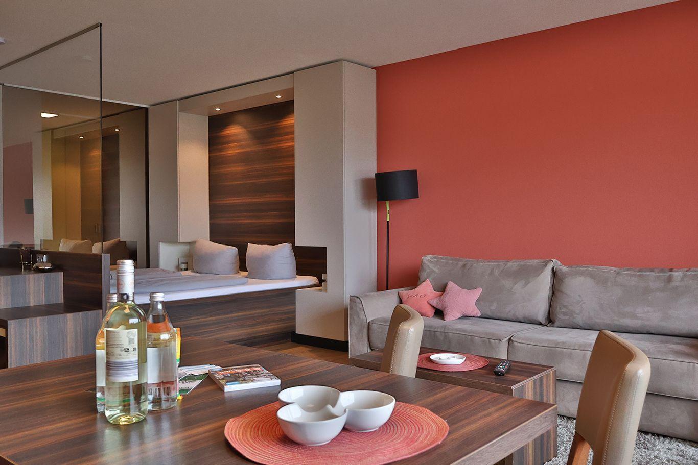 Wohnbereich mit Sofa, Tisch und Essbereich. Rote Wand, Spiegel und Bett im Hintergrund.