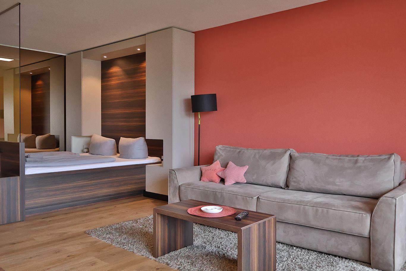 Ein Wohnbereich mit grauem Sofa, roter Wand und Holzboden. Im Hintergrund ist ein Bett sichtbar.