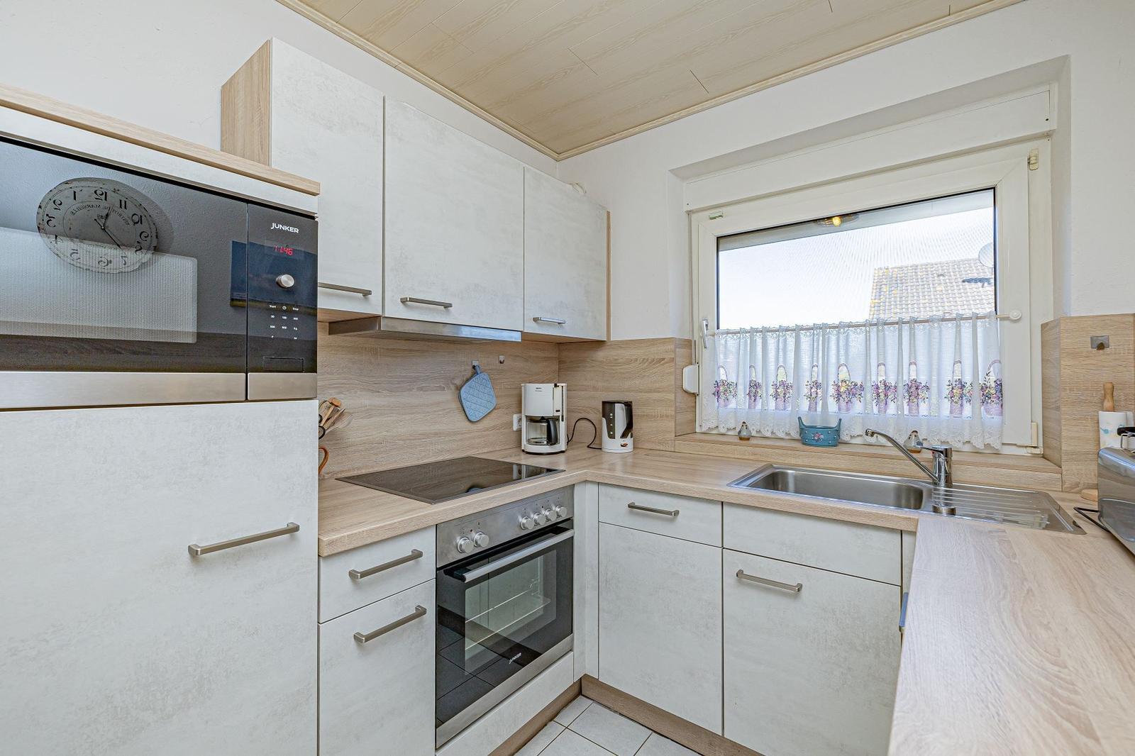 Küche mit weißen Schränken, Edelstahlspüle und Fenster mit Vorhang.