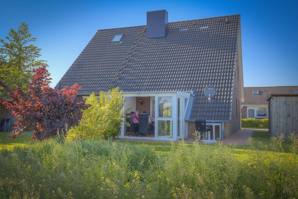Haus mit Glasveranda, Garten und Satellitenschüssel. Personen sitzen im Innenbereich.