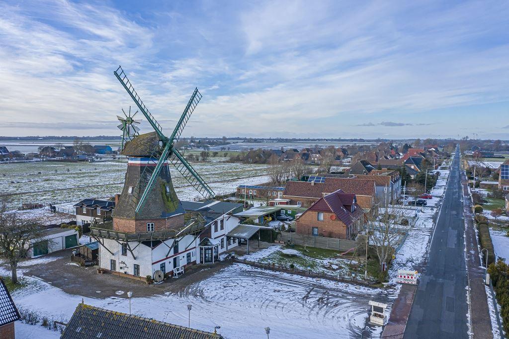 Schneebedeckter Ort mit historischem Windrad und Wohnhäusern entlang einer Straße.