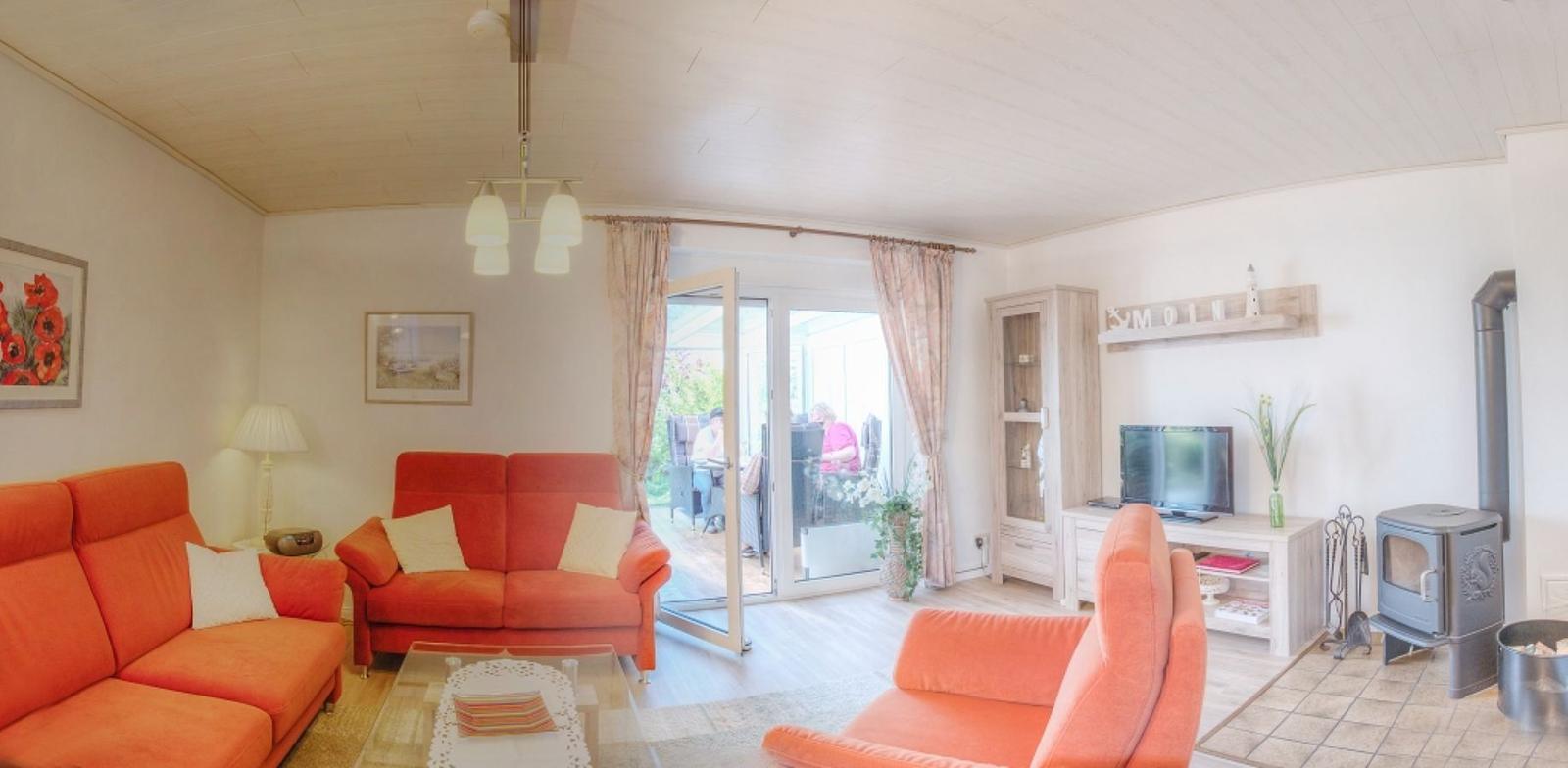 Wohnzimmer mit orangefarbenen Sofas, TV und Kaminofen. Durchgang zur Terrasse.