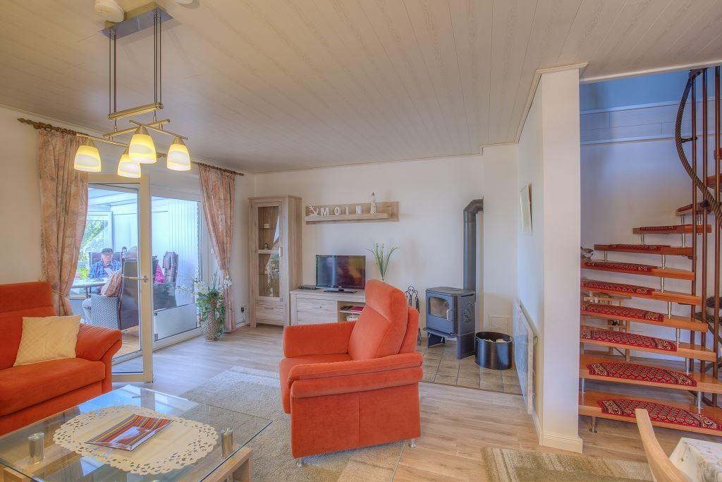 Wohnzimmer mit orangefarbenen Sitzgelegenheiten, Glas-Tisch und Holzboden. Treppe führt nach oben.