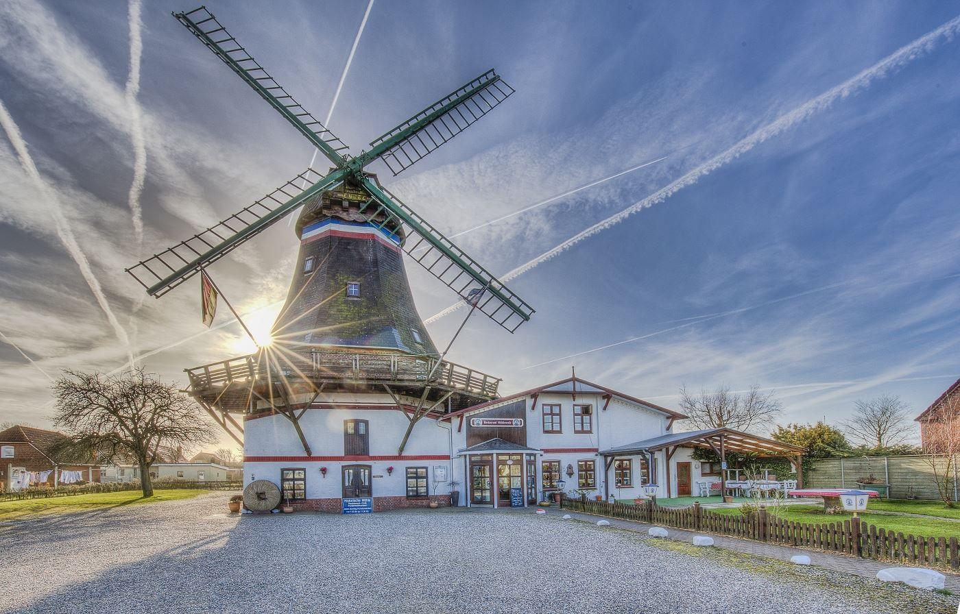 Windmühle mit Anbauhaus und Gartenbereich unter blauem Himmel mit Sonnenstrahlen.
