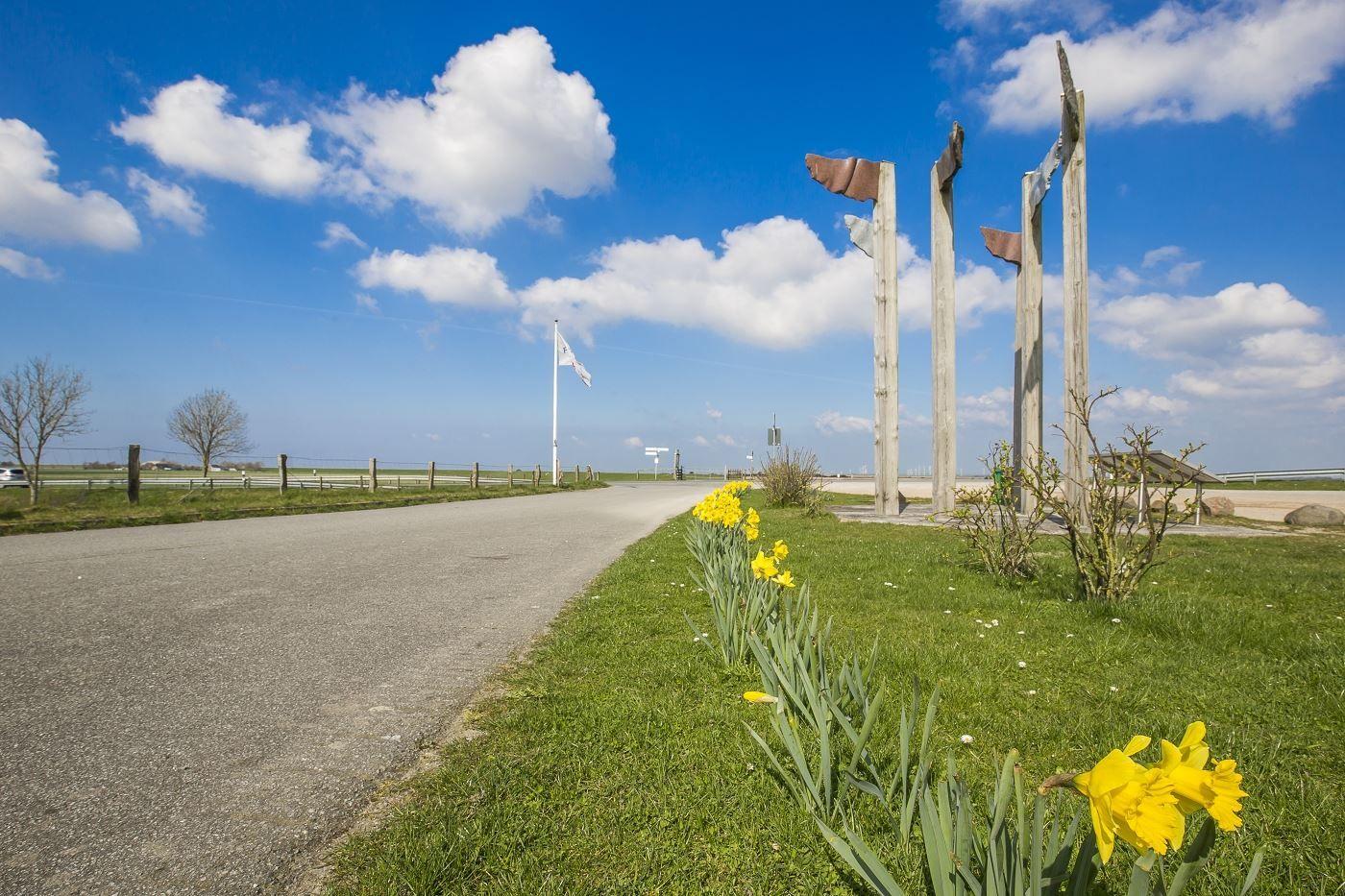 Eine Straße mit gelben Narzissen und einem Kunstwerk aus Holzpfosten unter blauem Himmel.
