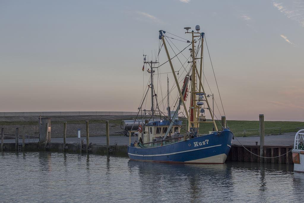 Ein blaues Fischerboot mit Masten liegt im Hafen. Der Himmel ist rosa und blau.