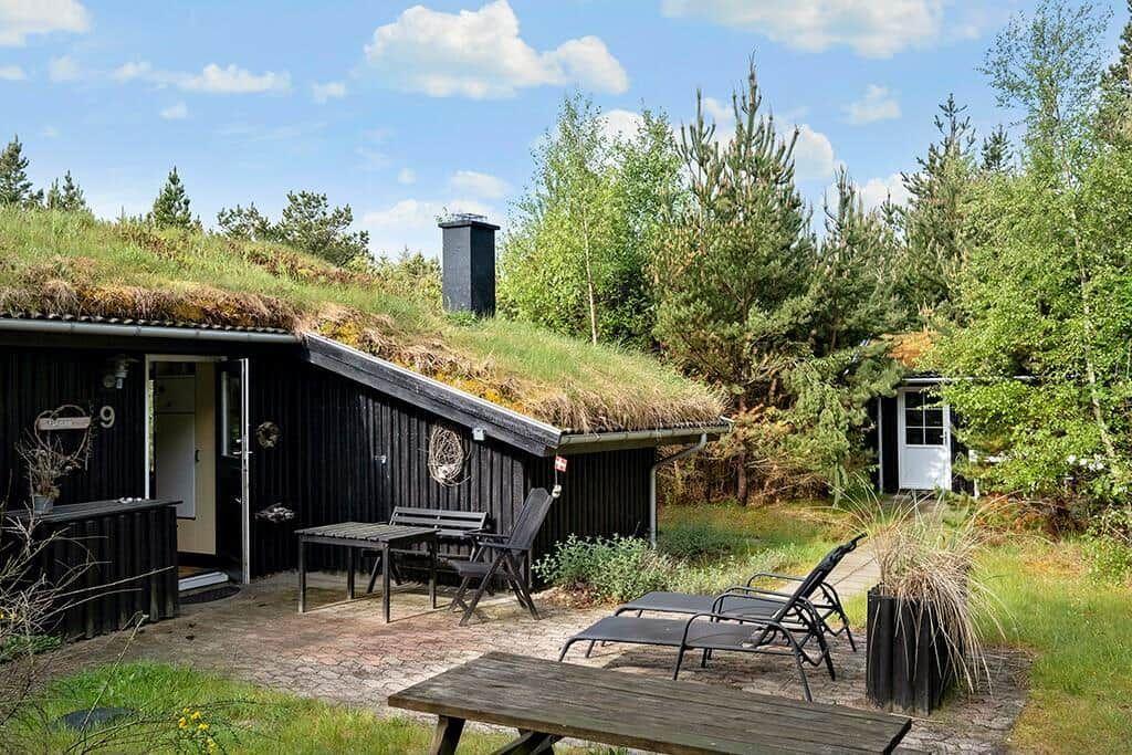 Haus mit grüner Dachdeckung, Terrasse mit Tisch, Stühlen und Liegen. Umgeben von Bäumen.