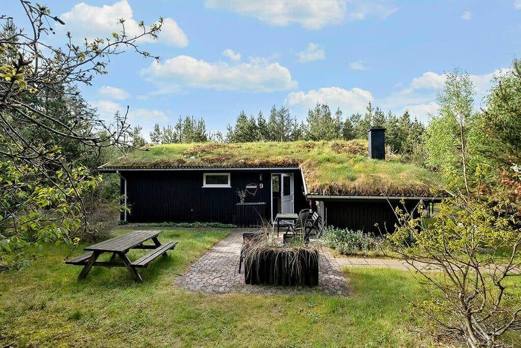 Ein dunkles Holzhaus mit grünem Dach, Garten und Picknicktisch unter blauem Himmel.