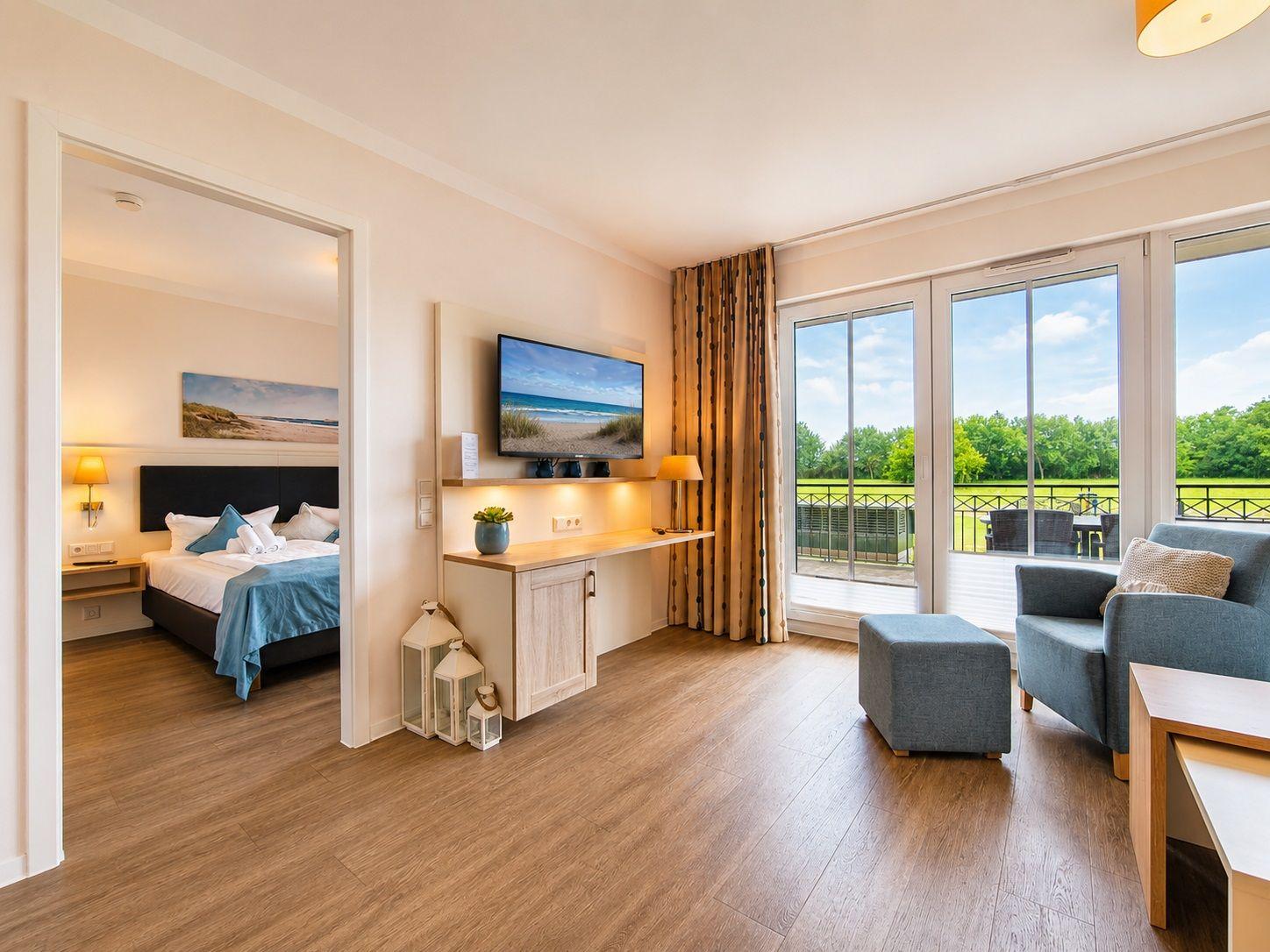 Schlafzimmer mit Schreibtisch, Fernseher und Fenster. Bett sichtbar im Hintergrund.