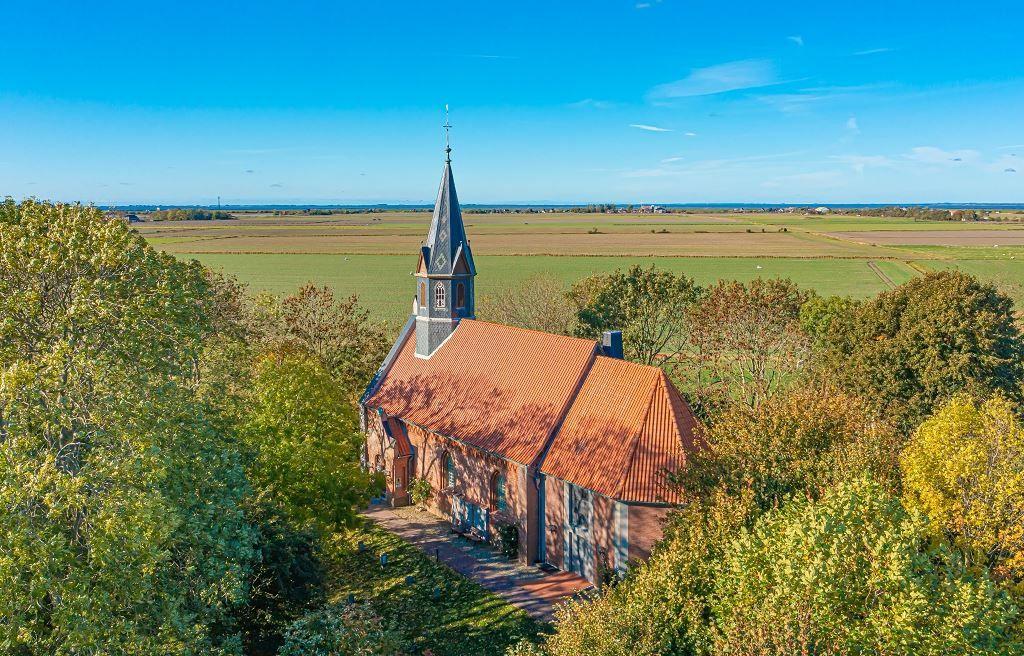 Kirche mit Turm und rotem Dach umgeben von Bäumen und Feldern unter blauem Himmel.
