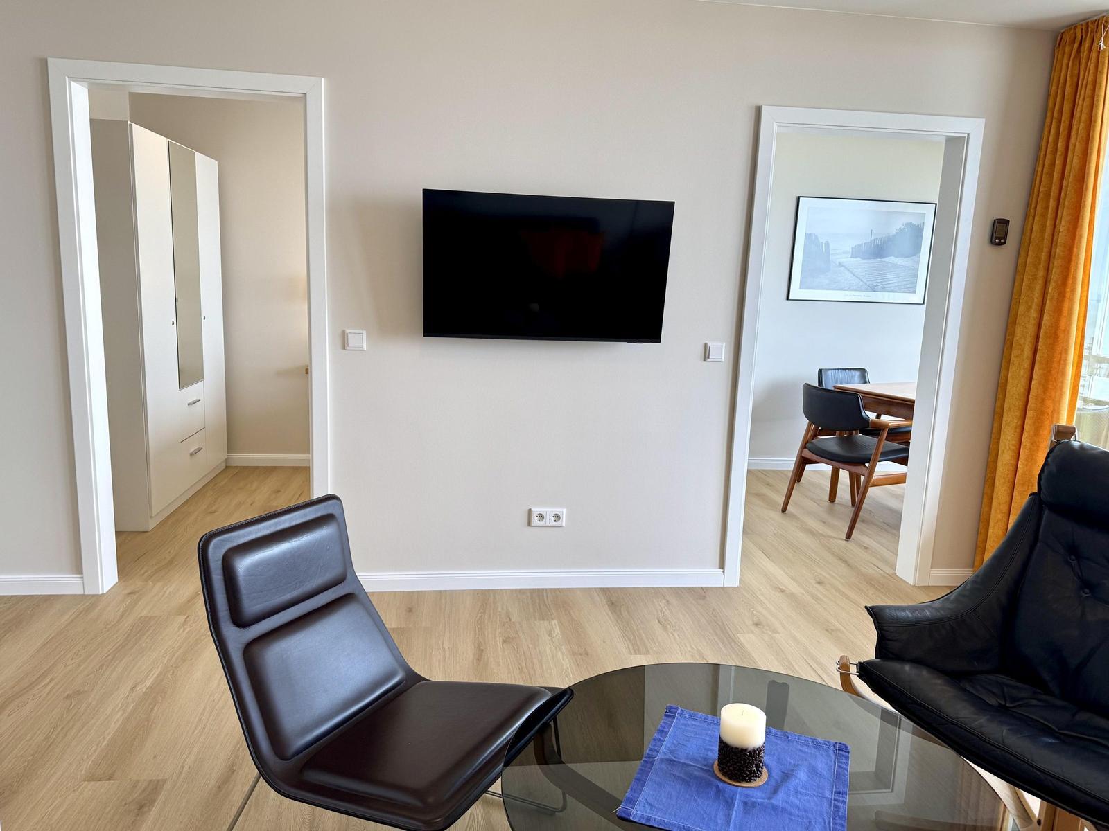 Living room with TV, two black chairs, and glass table. Doorways to bedroom and dining area.