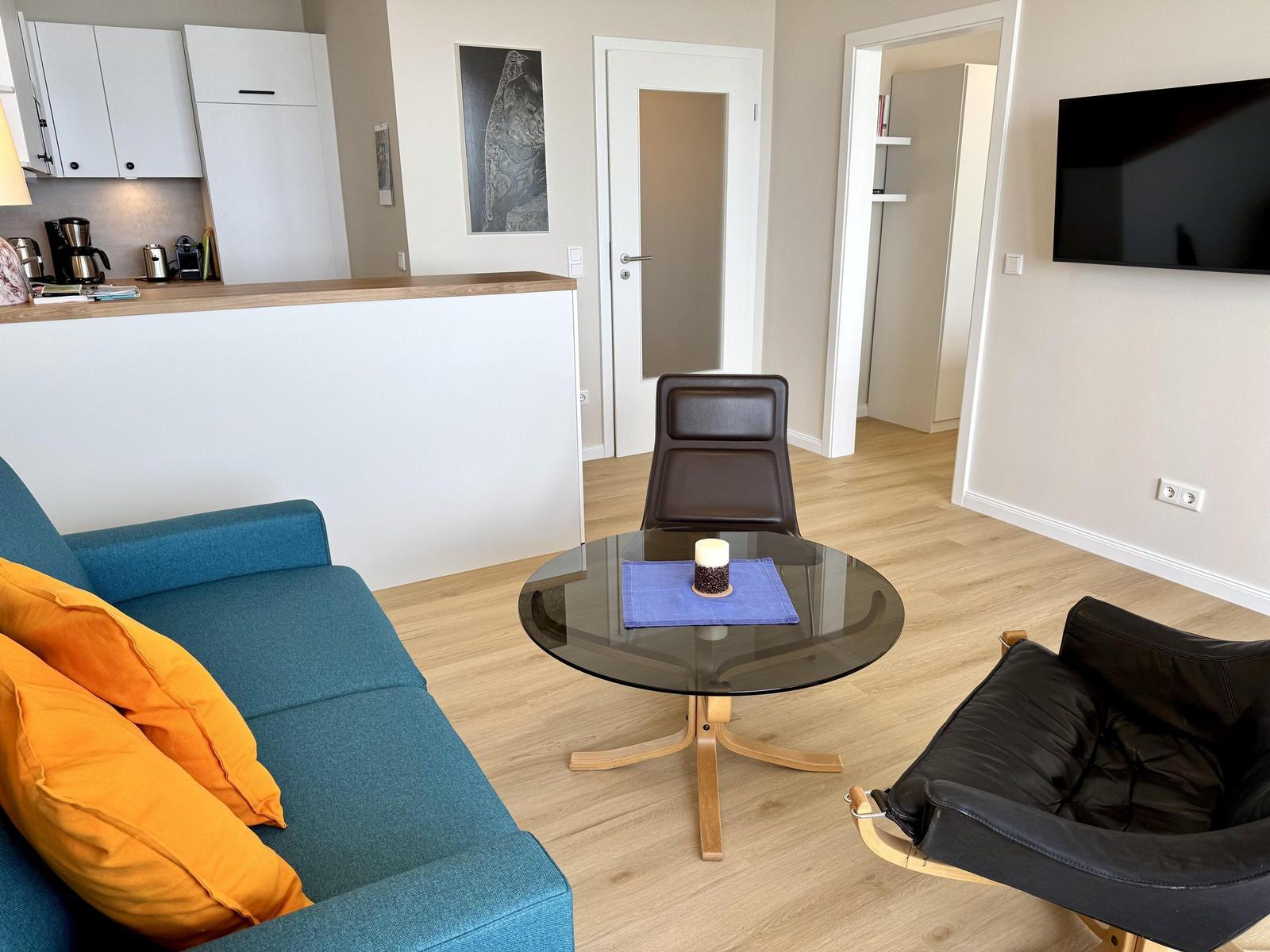 Living room with sofa, armchair, and table. Kitchen and TV are visible.