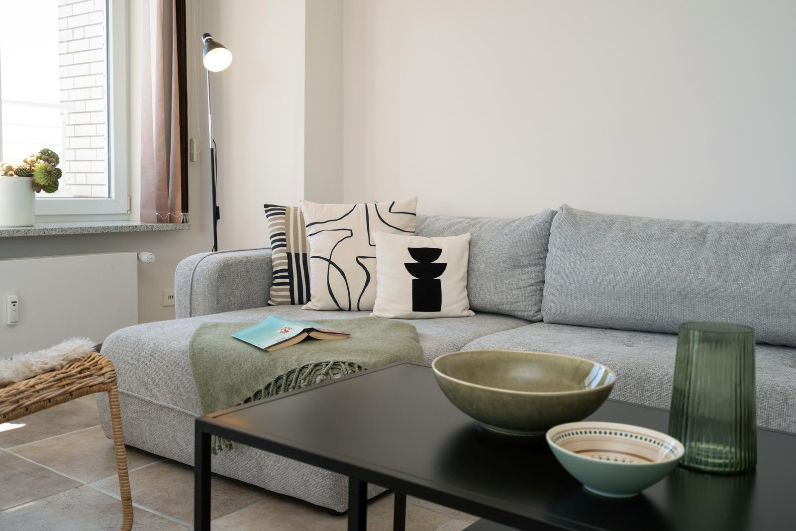 Ein grauer Sofa mit Kissen und einer Decke. Auf dem Tisch stehen Schalen und ein Glas.