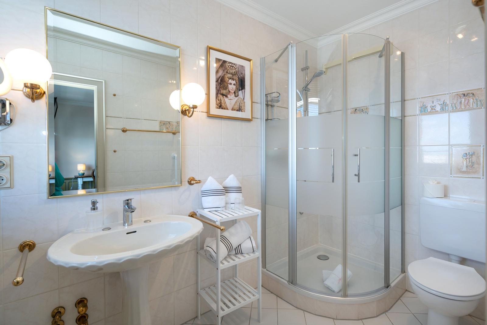 Badezimmer mit Dusche, Waschbecken und Toilette. Weiße Fliesen und goldene Accessoires.
