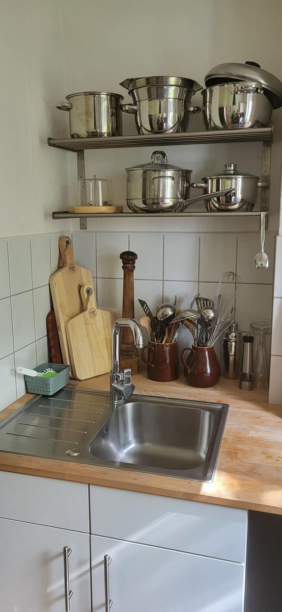 Küchenbereich mit Edelstahlspüle, Holzoberfläche und Metallregalen mit Kochgeschirr.