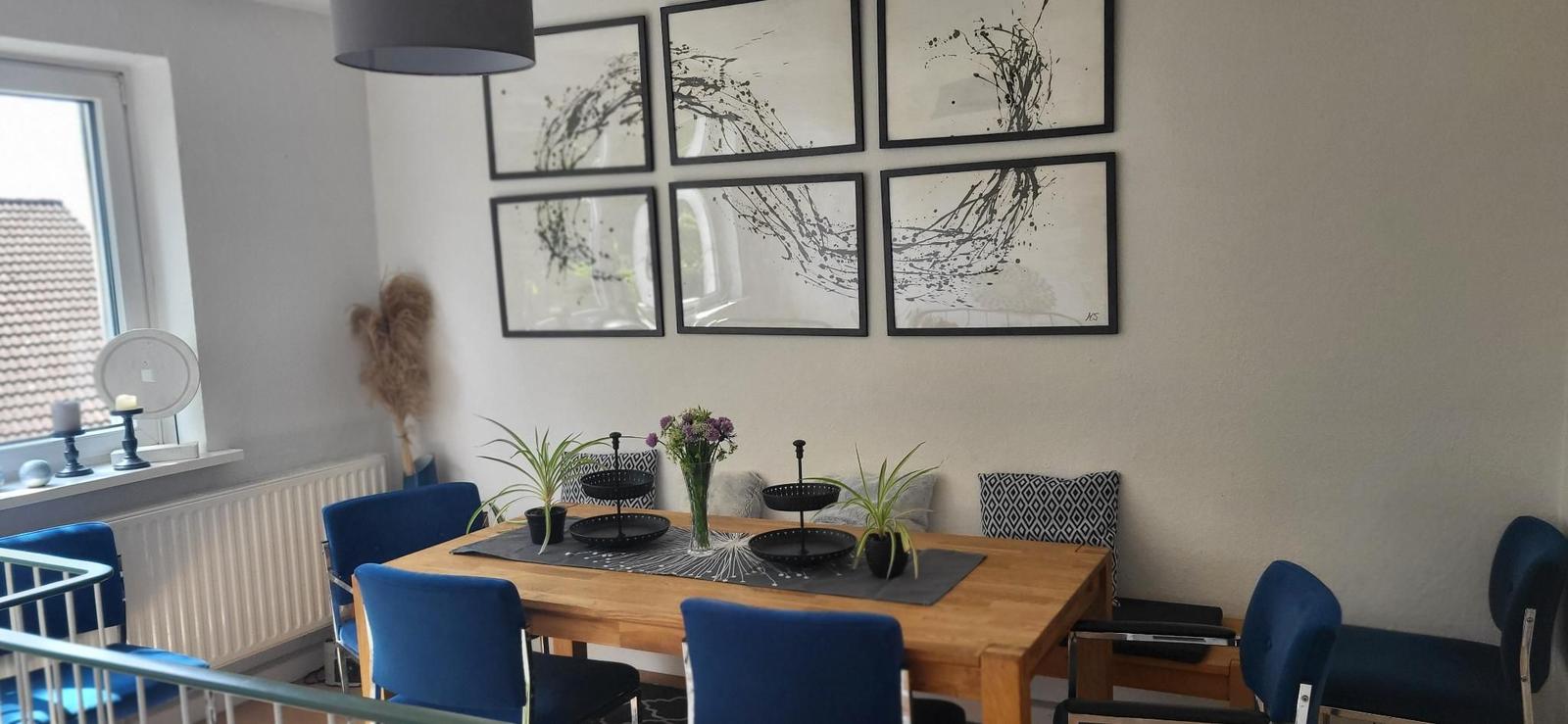 Esszimmer mit Holztisch, blauen Stühlen und sechs Wandbildern.