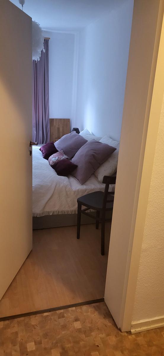 Schlafzimmer mit Bett, Kissen und Stuhl. Holzboden und Fenster mit Vorhängen.