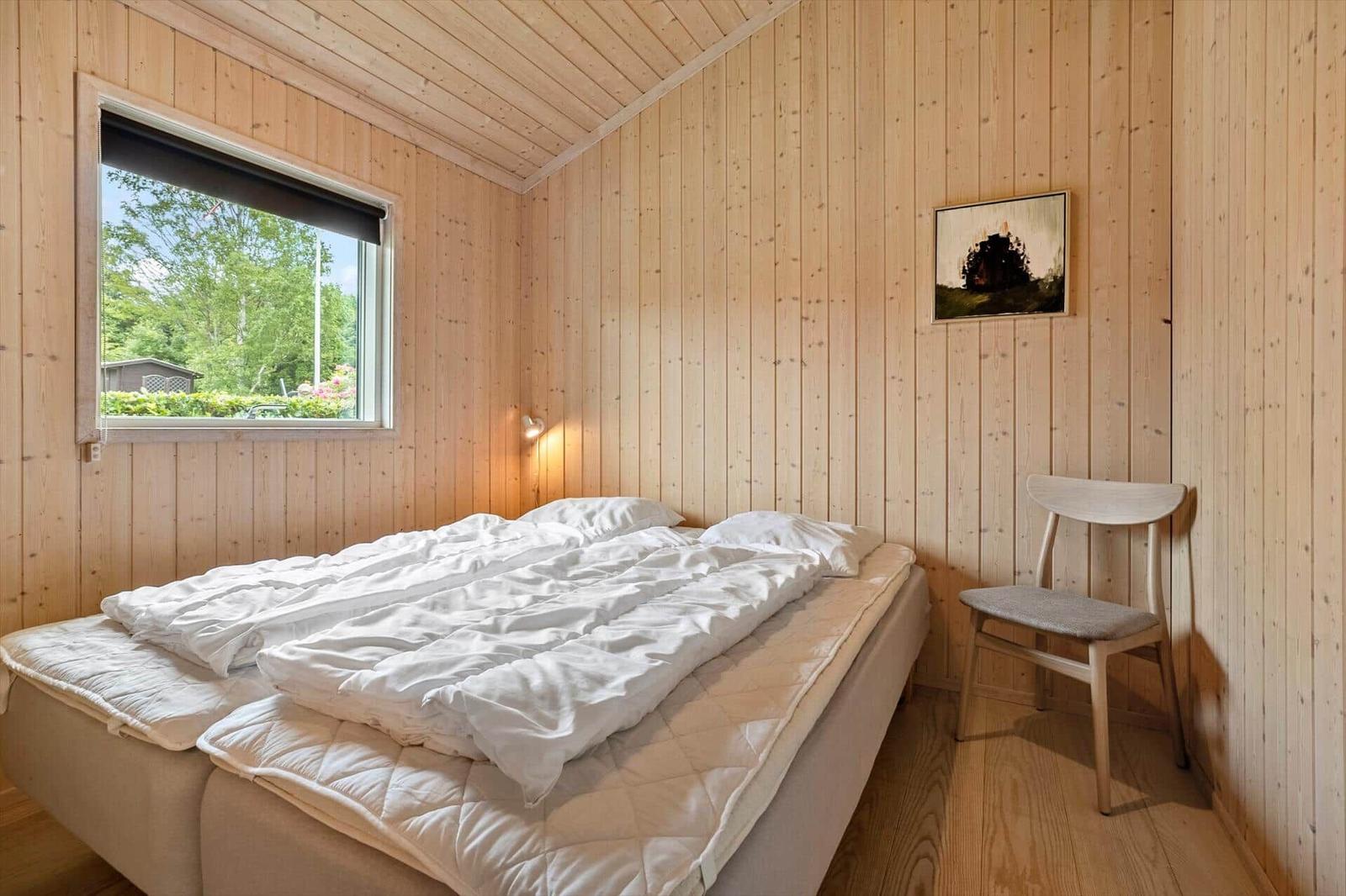 Bedroom with wooden walls, bed, chair, and window with view of greenery.