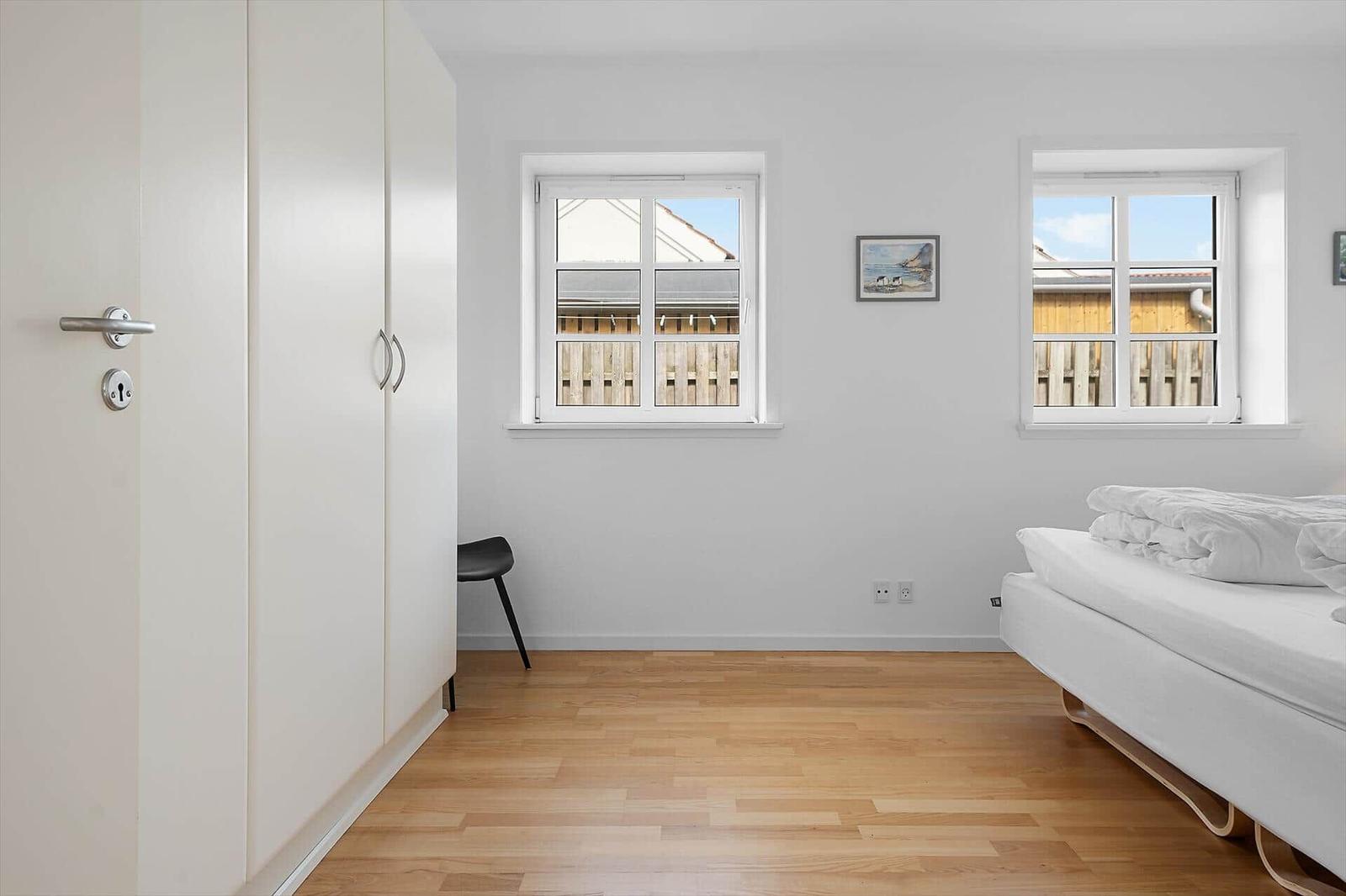 Schlafzimmer mit weißem Bett, Holzboden und zwei Fenstern. Links steht ein weißer Schrank.